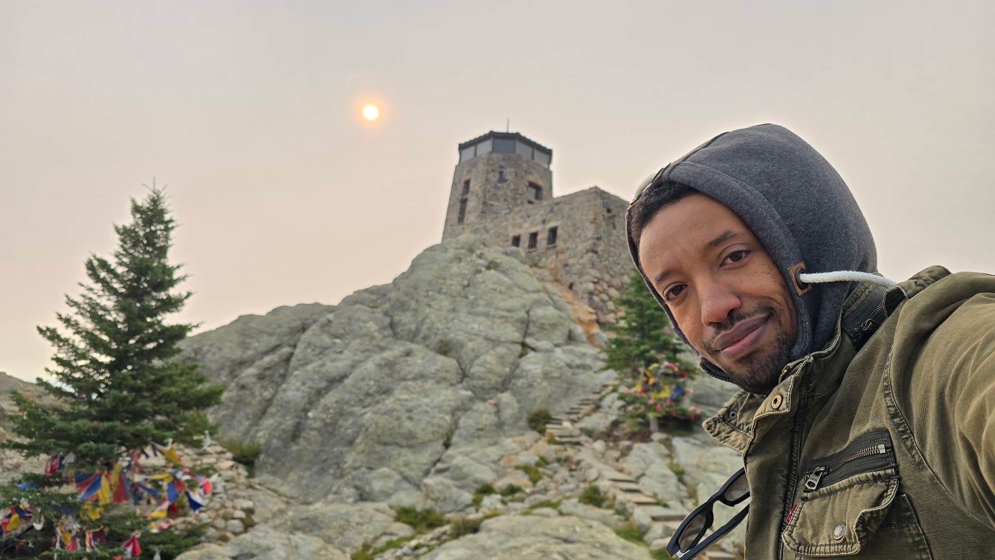 Yad in a grey hood and an olive jacket, close to the camera at the top of a rocky peak, a stone lookout tower on the summit behind him and a low sun through hazy sky.