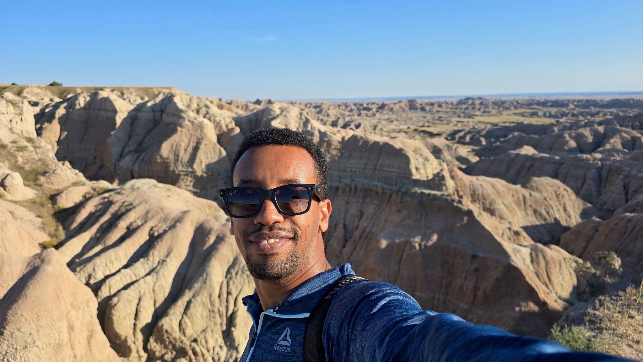 Yad in sunglasses and a blue quarter-zip, smiling at arm's length on the rim of a canyon, pale layered rock formations and open plains stretching out behind him.