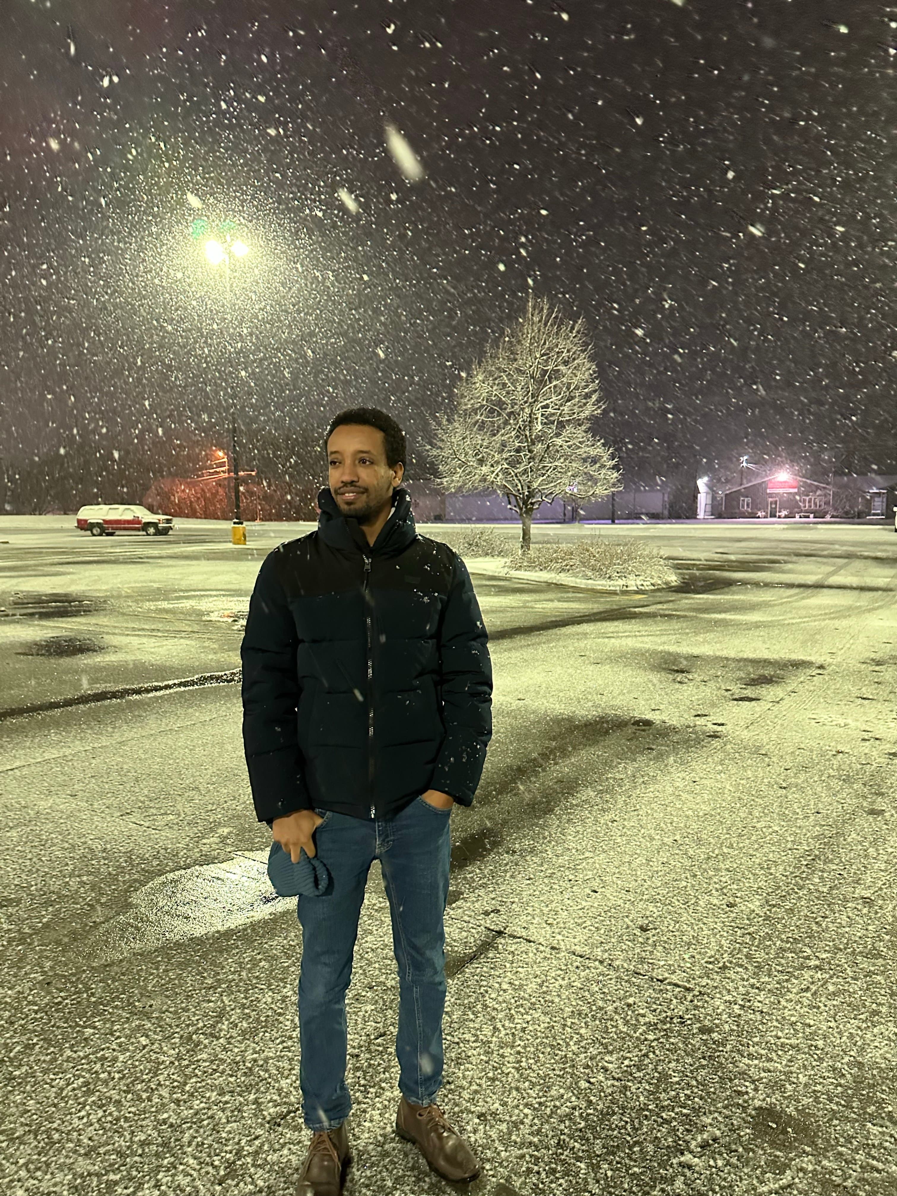 Yad standing in the falling snow at night, hands in his pockets, a lit tree and a quiet street behind him.
