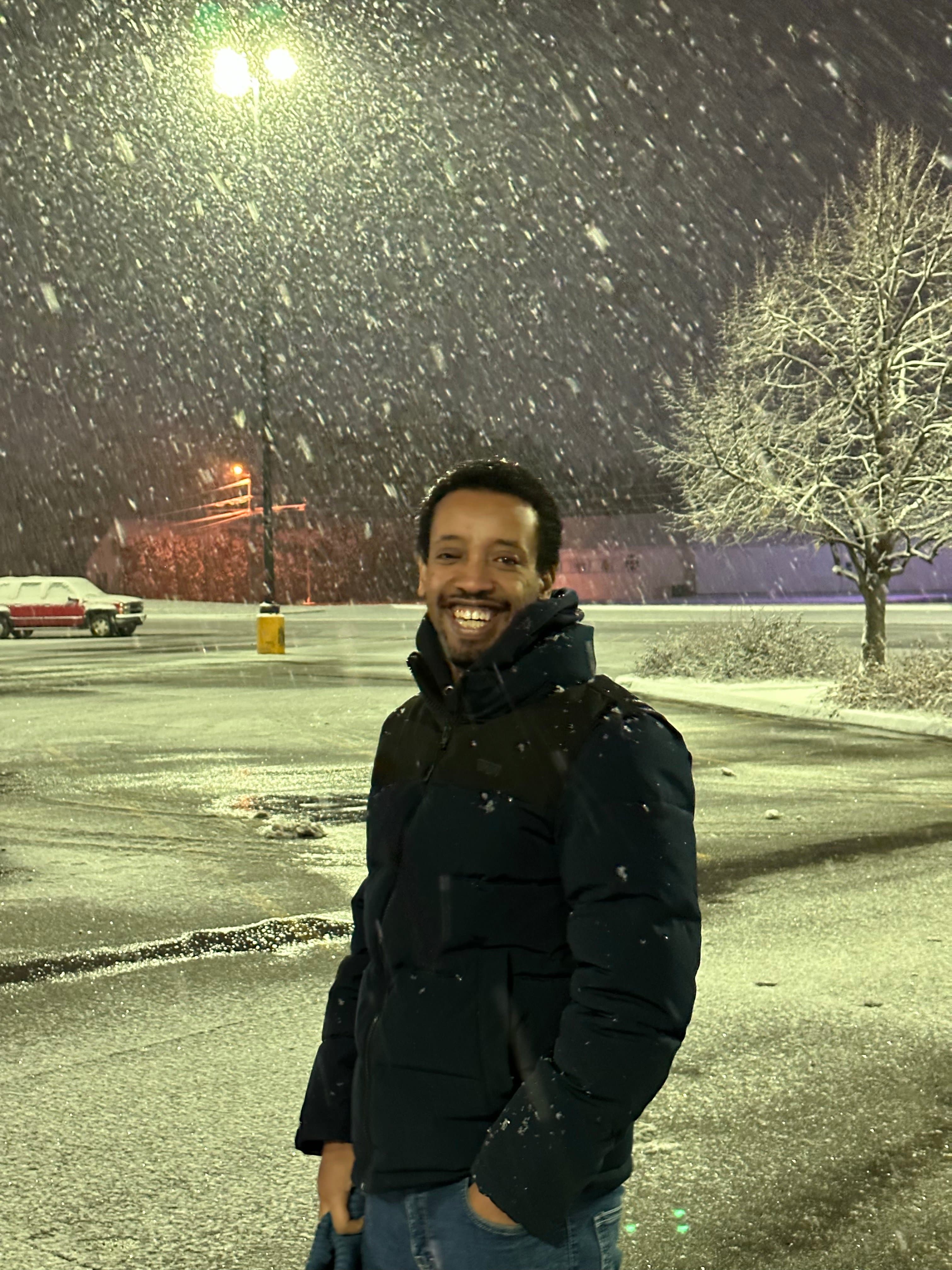 Yad laughing in a black puffer jacket in an empty parking lot at night, snow falling hard through the streetlight.