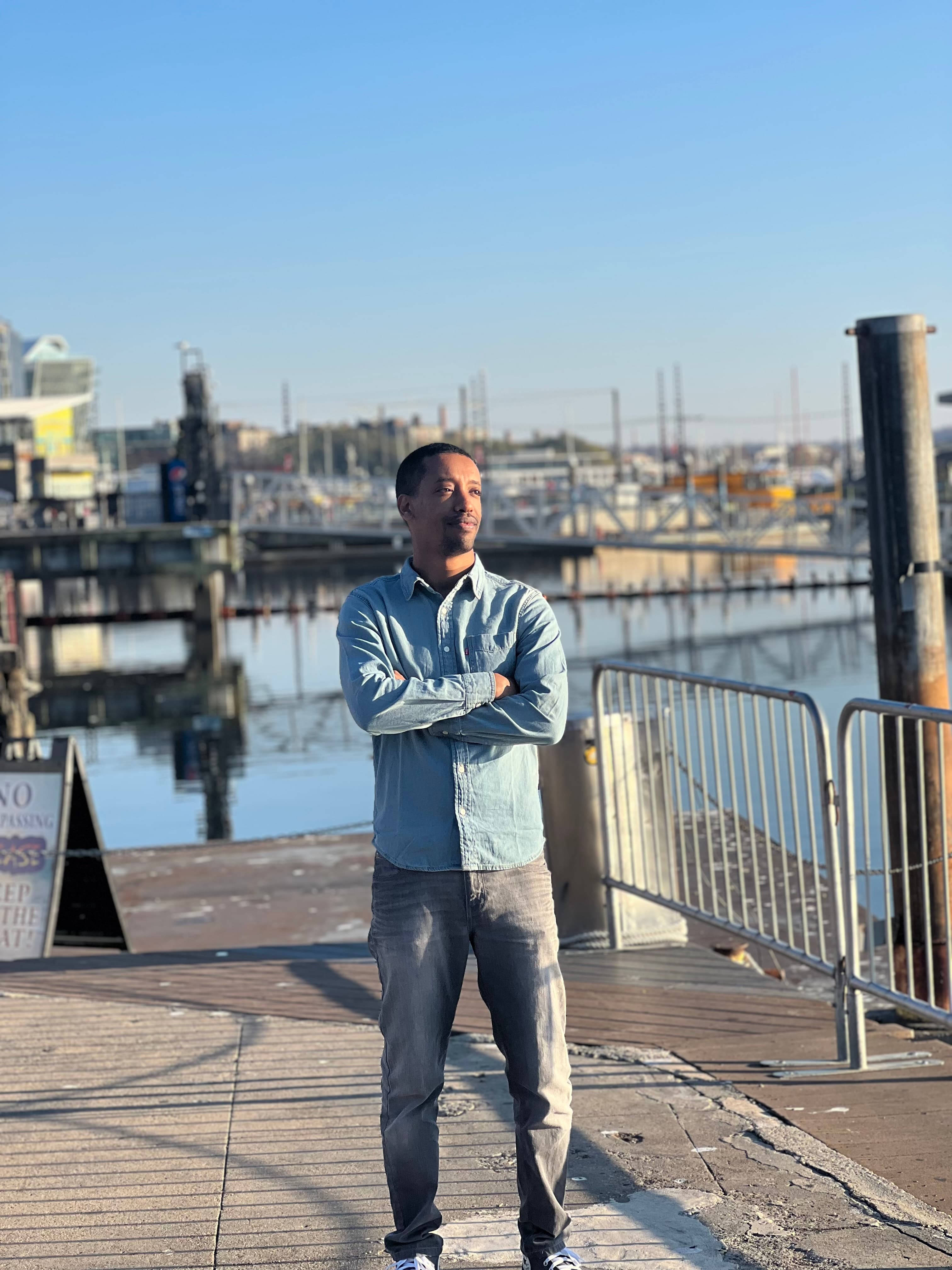 Yad on a waterfront boardwalk in a denim shirt, arms folded, looking off toward the marina on a clear blue day.