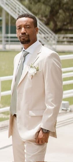 Yad in a cream suit with a white rose boutonnière and a pale green tie, standing on the grass at a wedding.