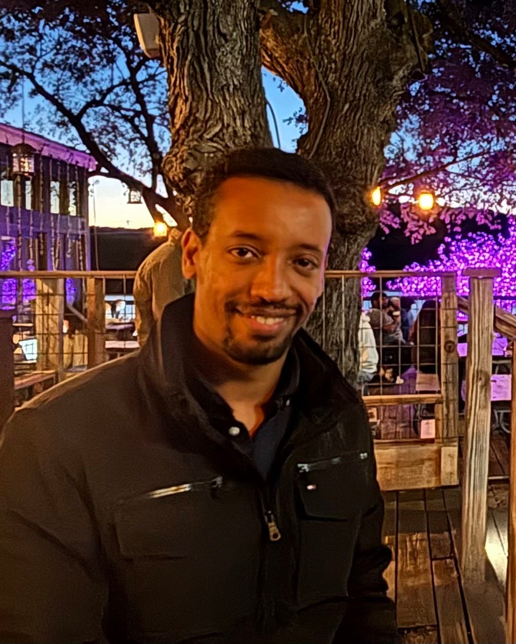 Yad smiling at a table under a big tree strung with lights, purple and gold evening light behind him.