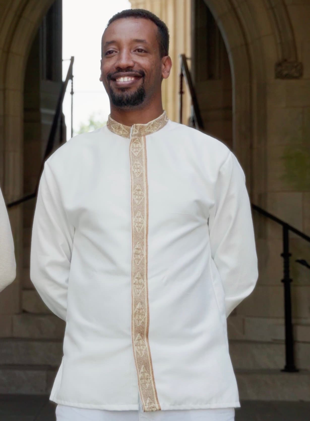 Yad in a white and gold embroidered shirt, beaming, on the stone steps of a church.