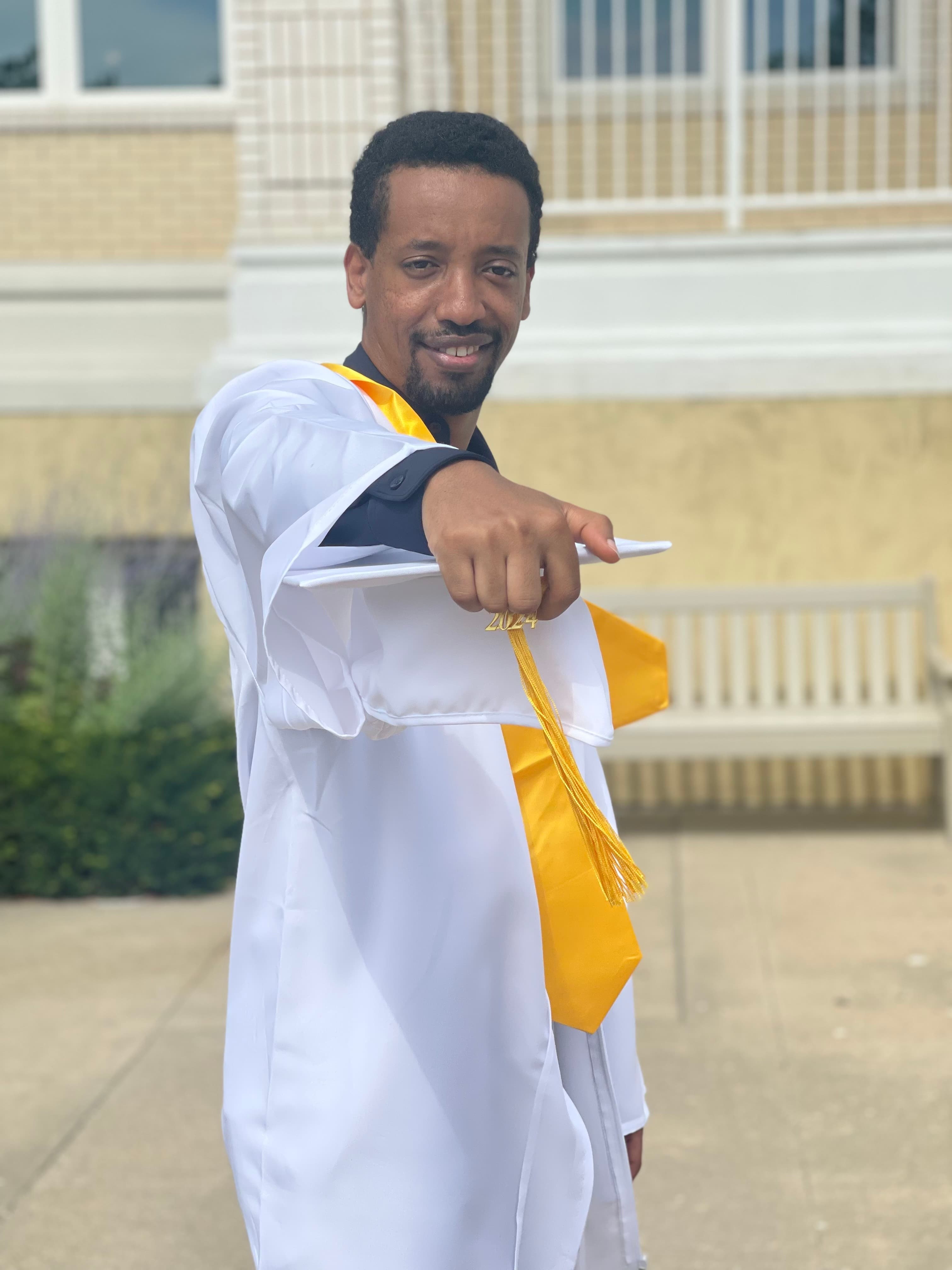 Yad in a white graduation gown and gold stole, holding his cap out toward the camera with a wide smile.