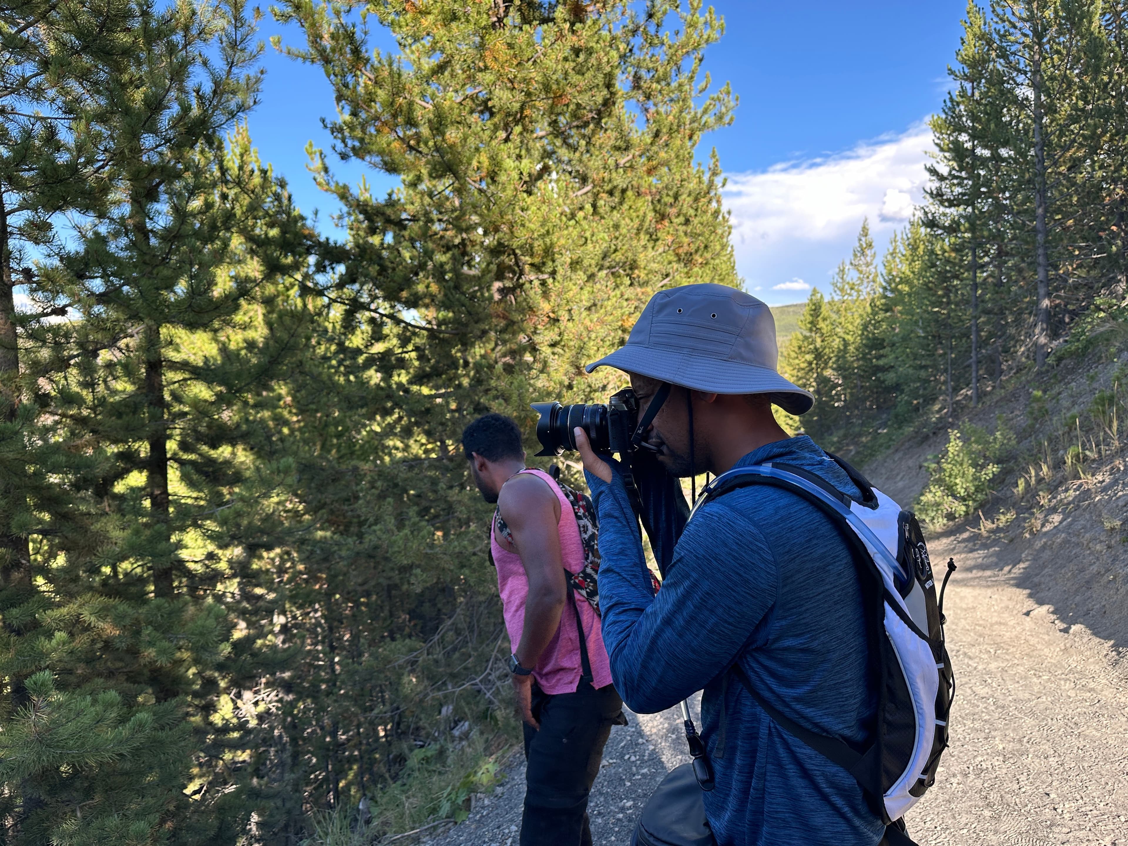 Yad in a bucket hat and backpack, raising his camera to his eye on a pine-lined mountain trail.