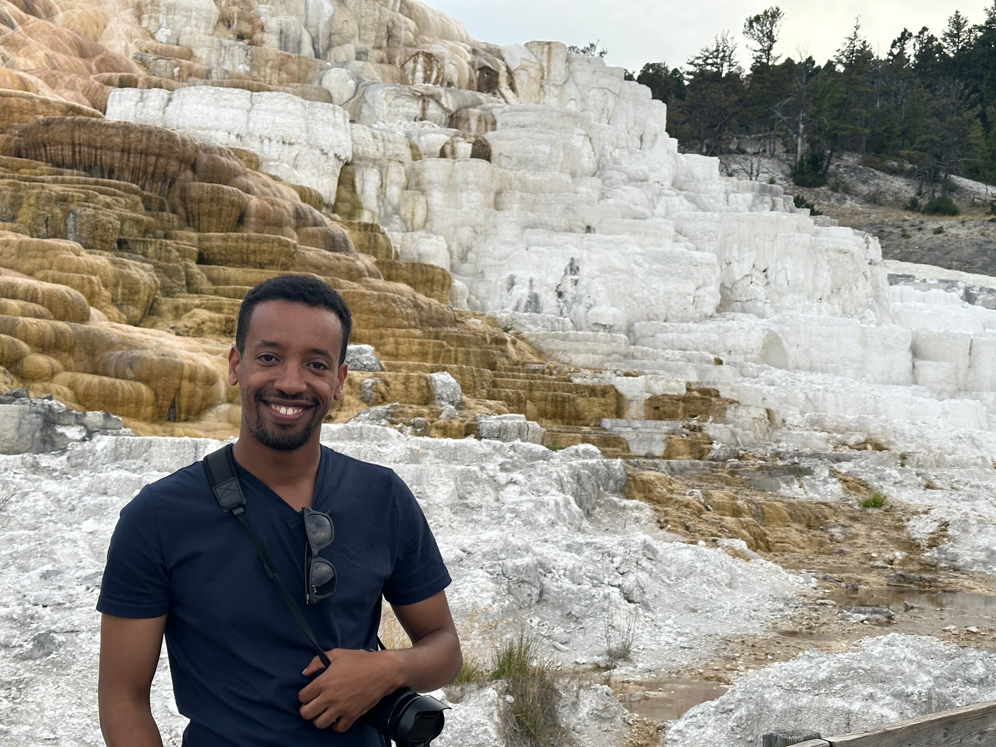 Yad grinning in a navy t-shirt with a camera slung at his side, in front of the white and ochre limestone terraces of a hot spring.