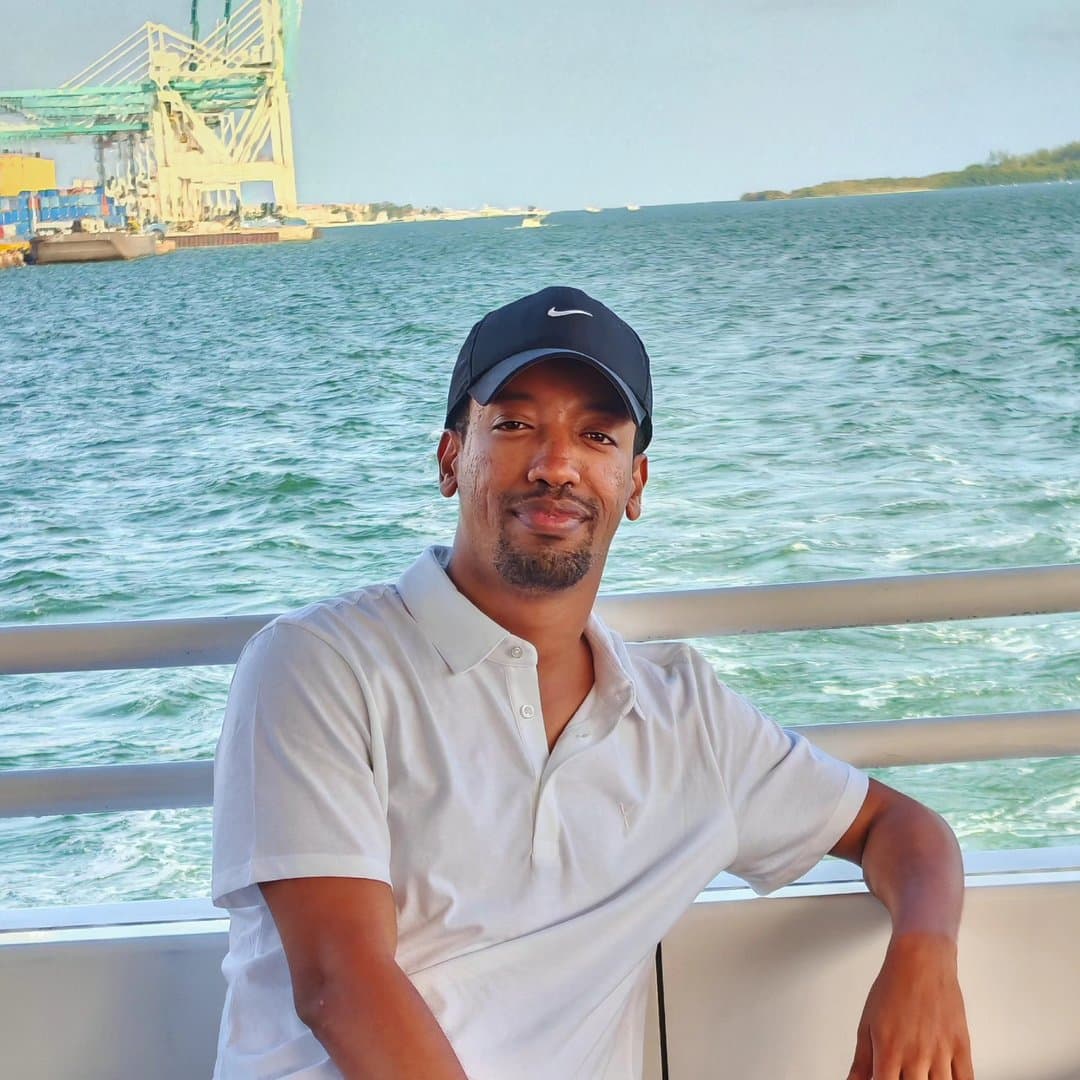 Yad in a white polo and a black cap, leaning back against the rail of a boat, green harbor water and shipping cranes behind him.
