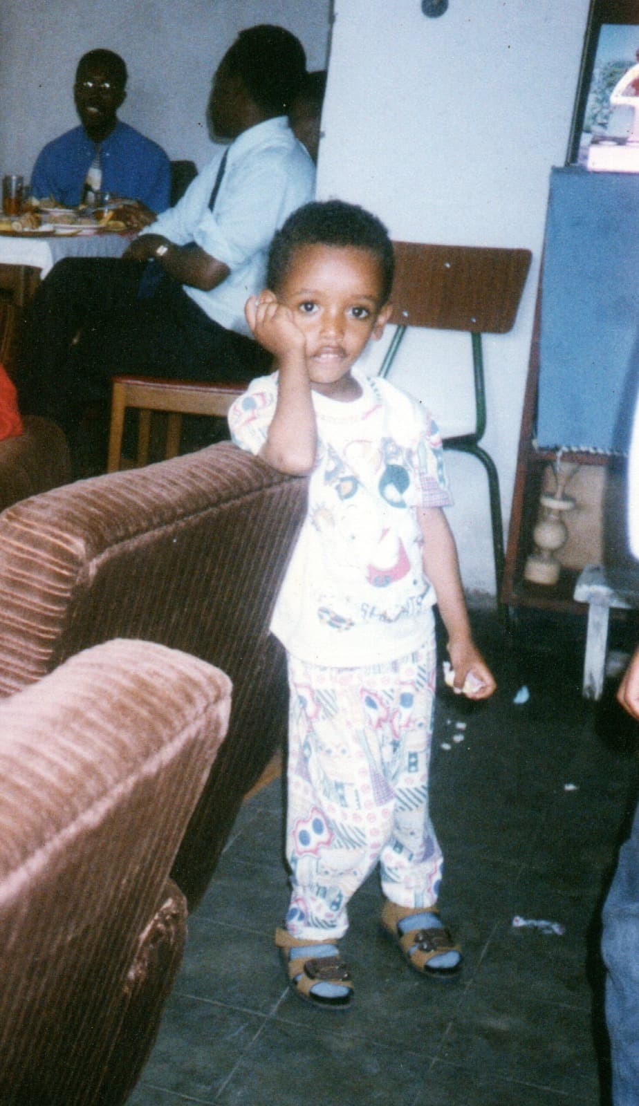 Yad as a toddler in a patterned pyjama set and small sandals, holding onto the arm of a velvet sofa and looking at the camera, adults sitting at a table behind him.