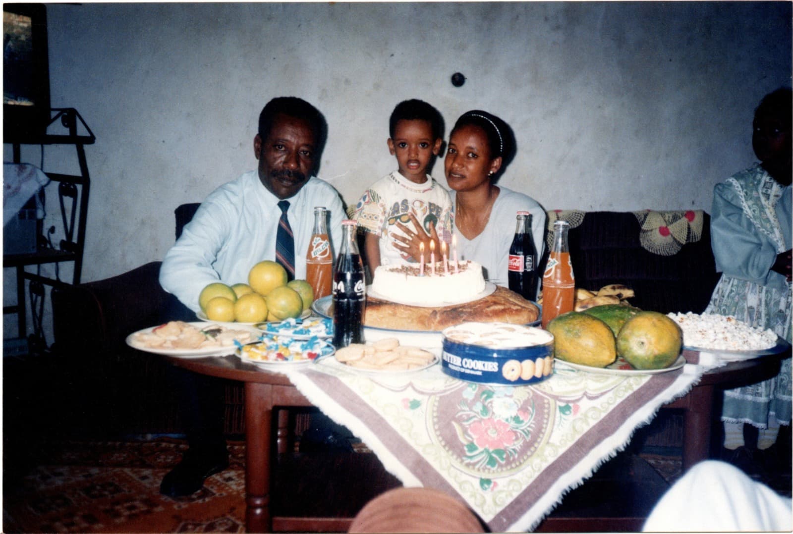 Yad as a small boy standing between a man in a shirt and tie and a woman in pale blue, behind a birthday cake with lit candles on a table crowded with fruit, bottles and plates of biscuits.