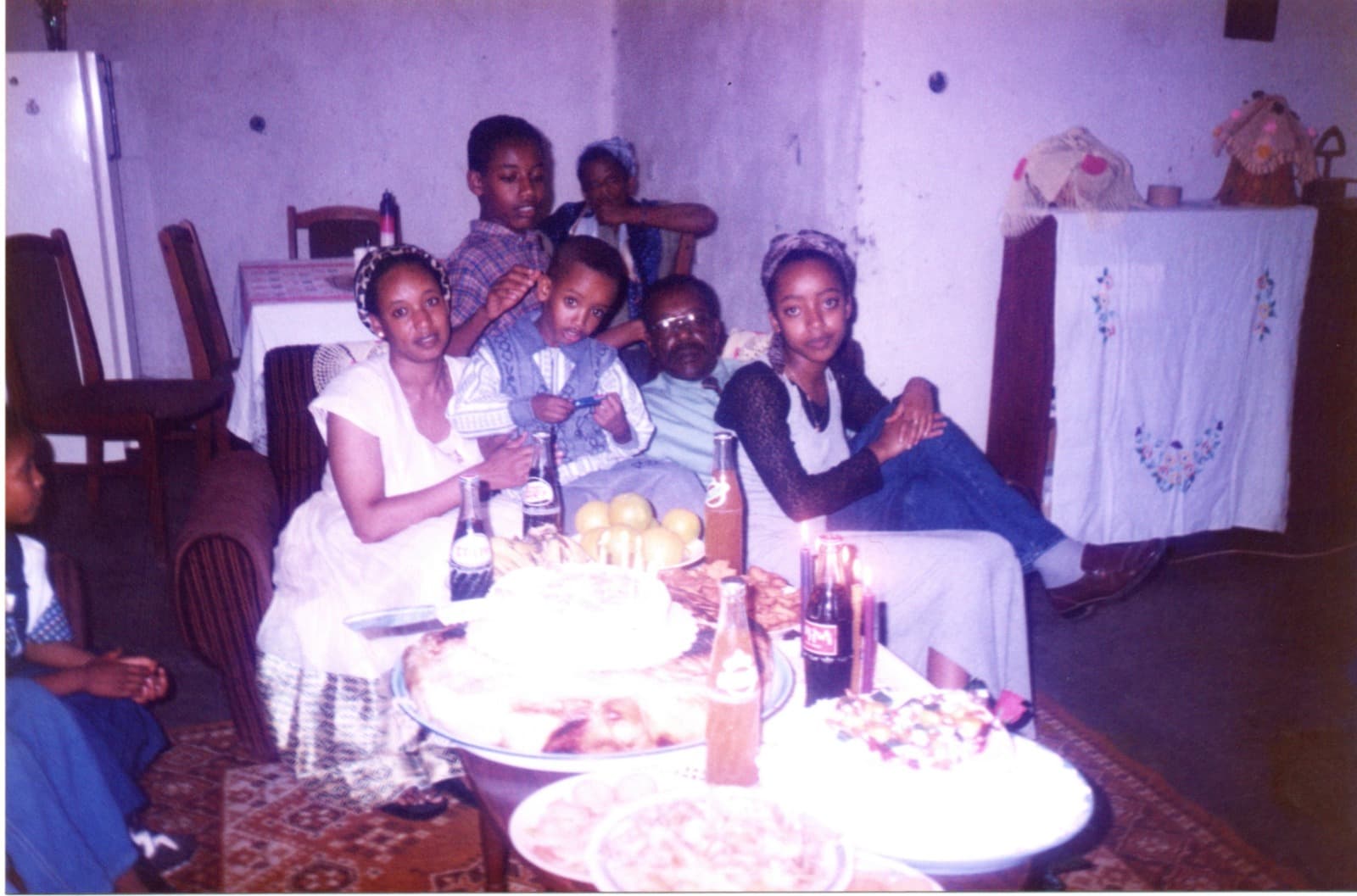 A family gathered on a sofa around a low table of cake and bottles, a woman in white holding a small boy on her lap, an older man in glasses and several children beside them.