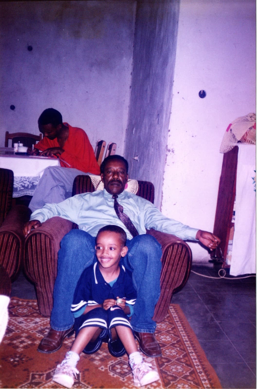 Yad, about five, in a navy tracksuit and white trainers, sitting on a patterned rug between the feet of a man reclining in an armchair in a shirt and tie.