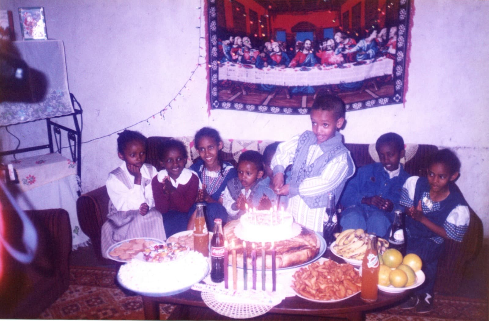 A room full of children on a sofa watching a boy lean in to cut a birthday cake ringed with lit candles, a Last Supper tapestry and a string of lights on the wall behind them.
