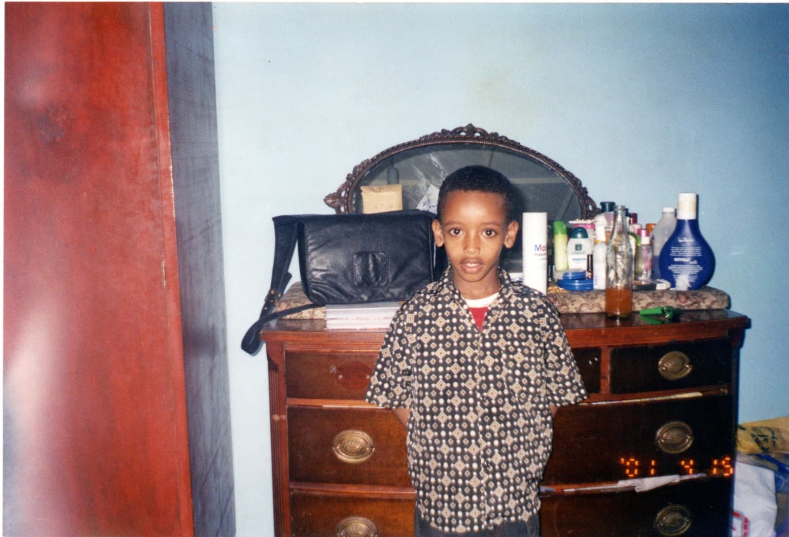 Yad as a boy in a patterned short-sleeved shirt, standing on his own in front of a wooden dresser with an oval mirror, against a pale blue wall.