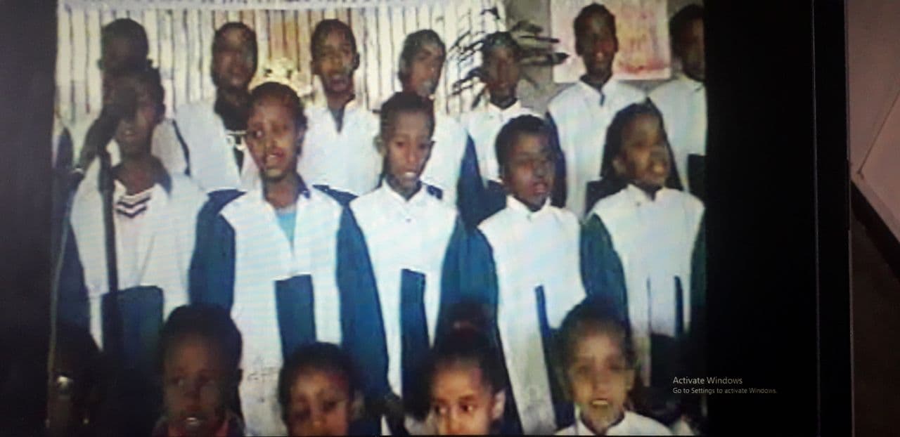 A children's choir in white shirts and blue robes singing in rows, photographed from a screen, so the picture is soft and grainy.